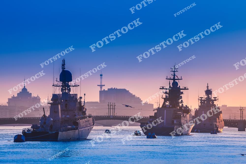 На фотографии запечатлены военные корабли, стоящие на Неве в Санкт-Петербурге в утренние часы. Небо окрашено в нежные пастельные тона рассвета, что создаёт атмосферу спокойствия. На заднем плане видны очертания городских зданий и мостов, добавляющих городской пейзаж к композиции. Плавные очертания судов на фоне восходящего солнца придают снимку особое настроение и величественность.